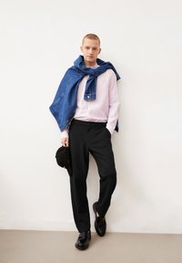 Lichtroze blouse met lange mouwen, zwarte op maat gemaakte broek, spijkerjasje over de schouders, zwarte pet in de hand en zwarte schoenen.