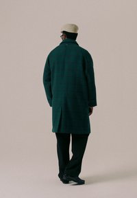 Grønn rutete ullfrakk med avslappet passform, frontknapper og sidelommer, kombinert med svarte bukser og en beige caps.