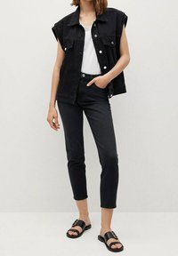 Chaleco sin mangas de mezclilla negra sobre una camiseta blanca, combinado con jeans oscuros y sandalias negras con un acento dorado. Diseño simple y moderno.