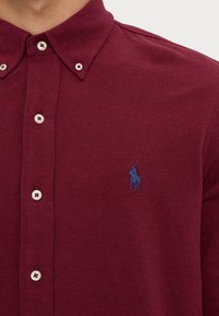 Polo color burdeos con tejido texturizado, cuello abotonado y botones redondeados. Presenta un pequeño logo bordado en azul en el pecho.