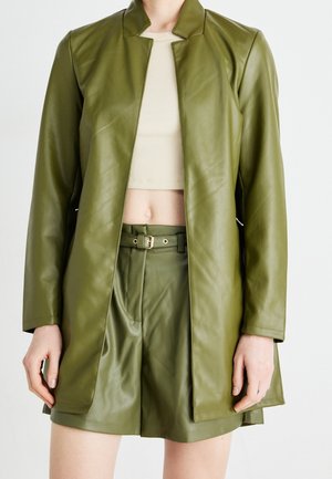 Lang jack van olijfgroen imitatieleren met lange mouwen en een riembroek, gedragen met een beige cropped geribbelde top op een persoon.