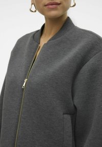 Pilkas zip-up bombonešis su nugarinėmis apykaklėmis, šoninėmis kišenėmis ir auksiniu užtrauktuku; pagamintas iš minkštos, tekstūruotos medžiagos.