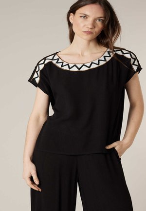 Femme portant un haut à manches courtes noir et un pantalon avec un bord en zigzag blanc et noir sur l'encolure et les épaules, posant avec la main dans la poche.