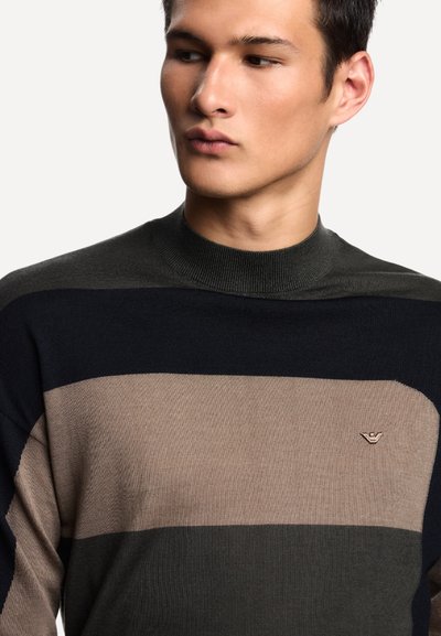 Giovane uomo che indossa un maglione a maglia a righe nei colori nero, beige e verde scuro, che guarda di lato su uno sfondo neutro.
