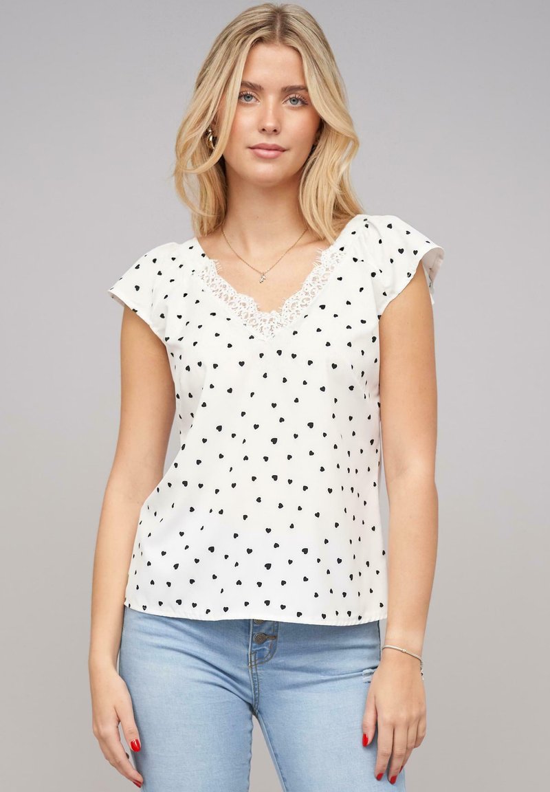 Blusa blanca de manga corta con escote en V, adornada con patrones de corazones negros y con detalles de encaje en el escote. Tejido suave y liso.