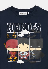 Námornícky modré tričko s vystylizovanými grafikami superhrdinov: Flash v červenej, Superman v svetlomodrej a Batman v tmavošedej. Zahrnuté sú aj mraky a netopiere.