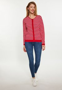 Czerwony i biały sweter w paski z dekoltem w serek, z zapięciem na guziki i ściągaczami przy mankietach, w połączeniu z niebieskimi jeansami skinny i białymi trampkami.