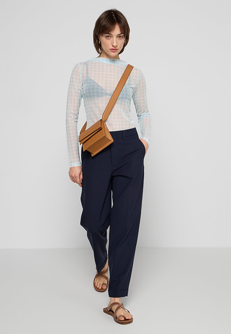 Helles blaues Langarmoberteil mit geometrischem Muster, kombiniert mit navyblauen Hosen und einer braunen Umhängetasche. Das Modell trägt flache Sandalen.