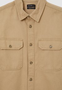 Beige Denimhemd mit Knopfleiste, spitzem Kragen, zwei Brusttaschen mit Klappen und kontrastierenden braunen Knöpfen. Reguläre Passform.