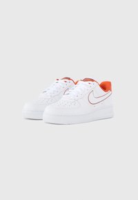 Biele tenisky Nike Air Force 1 s oranžovým akcentom na päte, vybavené perforovanou špičkou, plochými šnúrkami a textúrovanou gumovou podrážkou.