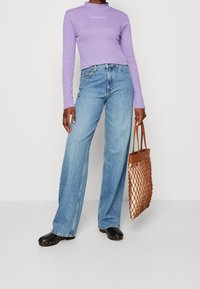 Fialový top s dlouhým rukávem a vysokým límcem, světle modré široké džíny, černé dřeváky a hnědá pletená taška přes rameno.