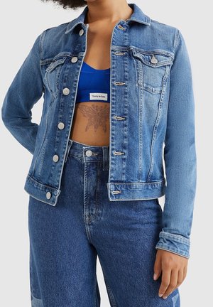 Person iført en blå denimjakke og jeans, med jakken åben, så en blå crop top og en sommerfugletatovering på maven kan ses.