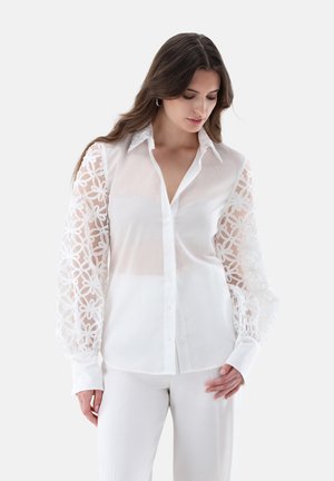 Woman wearing a white button-up blouse with sheer floral lace sleeves and white pants, looking down with long brown hair.