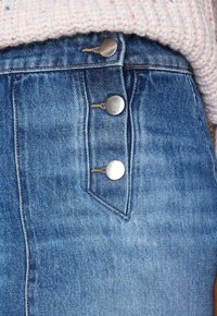 Närbild av en blå denimkjol med tre silverknappar och synlig beige sömnad, ihop med en ljusrosa stickad tröja.