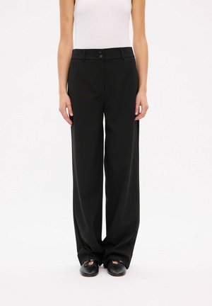 Femme portant un pantalon noir taille haute à jambes larges avec une fermeture à double bouton, associé à des ballerines noires et un haut blanc sans manches.