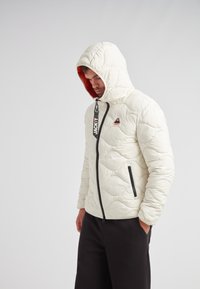 Puffjacka i off-white med ett kviltat mönster, med en huva i röd kant, dragkedjestängning och svarta detaljer på fickor och dragkedja.