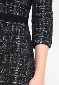 Svart och vit texturerad klänning med trekvartslånga ärmar och en solid svart midja. Tyget har ett prickigt mönster.