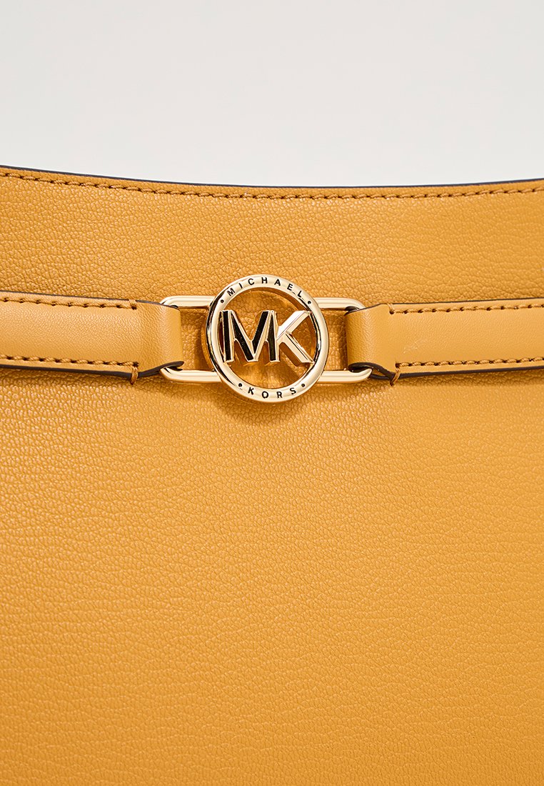 Gele leerachtige tas met een goudkleurig rond logo met "MK." Gladde schouderriem met gedetailleerde stikselaccenten.