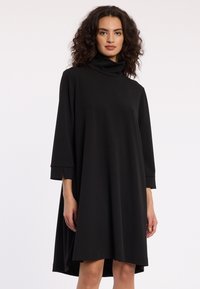 Vrouw met krullend haar die een losse zwarte coltrui-jurk met driekwart mouwen draagt tegen een effen witte achtergrond.