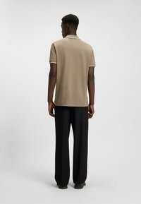 Långärmad, beige polo med vit kant, kombinerat med löst sittande svarta byxor, sedd bakifrån mot en enfärgad bakgrund.