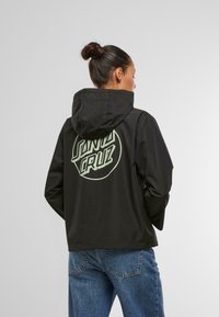 Black jacket with a hood, featuring a large mint green "SANTA CRUZ" logo on the back. Smooth texture and casual fit, worn with blue jeans.