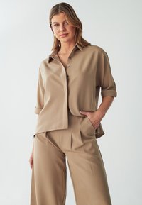 Beige button-up shirt with rolled sleeves, paired with matching high-waisted pleated trousers. Smooth fabric and relaxed fit.