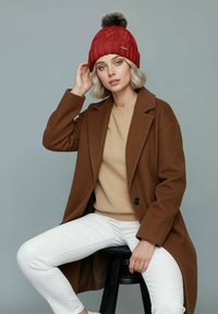 Red knitted beanie with a pom-pom, brown coat over a beige sweater, and white pants, seated on a black stool against a gray background.