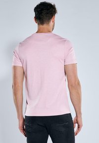 Hellrosa Kurzarm-T-Shirt aus weichem Stoff, mit rundem Ausschnitt und figurbetontem Schnitt. Trägt eine schwarze Hose und wird von hinten betrachtet.