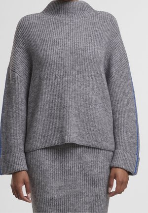 Personne portant un pull en grosse maille gris et une jupe assortie avec des rayures bleues sur les manches, debout les bras détendus le long du corps.