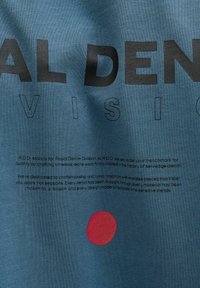 Zils audums ar izteiksmīgu melnu tekstu, kas stāsta "DENIM VISION", un sarkanu punktu. Teksts zemāk apraksta zīmola apņemšanos nodrošināt kvalitatīvu meistarību.