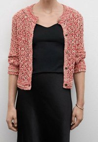 Pletený korálový cardigan s kulatým výstřihem, otevřeným pletením a stříbrnými knoflíky. Nosený přes černé šaty s dlouhými rukávy.