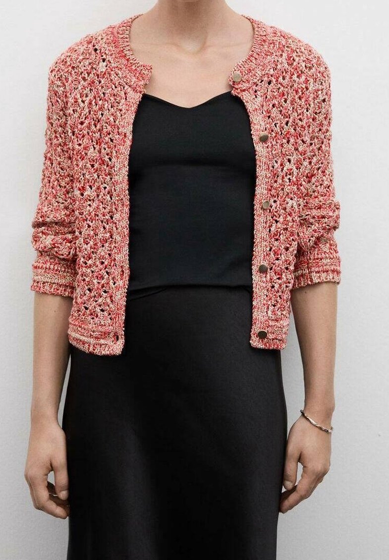 Pletený korálový cardigan s kulatým výstřihem, otevřeným pletením a stříbrnými knoflíky. Nosený přes černé šaty s dlouhými rukávy.