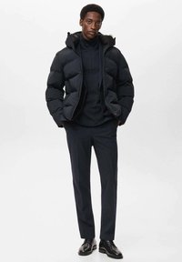 Giacca a vento nera con cappuccio, caratterizzata da sezioni trapuntate. Indossata sopra un maglione scuro e abbinata a pantaloni scuri su misura e scarpe nere.