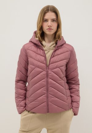 Jeune personne portant une veste matelassée mauve à capuche par-dessus un sweat à capuche beige, les mains dans les poches d'un pantalon beige, fond neutre.