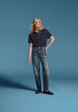 Femme souriante aux longs cheveux blonds portant un polo bleu marine, un jean bleu et des chaussures noires, debout devant un fond bleu uni.