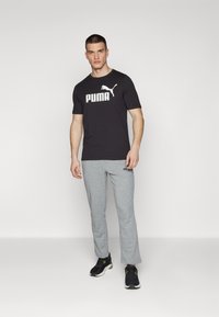 Melna kokvilnas T-krekls ar izteiksmīgu baltu "PUMA" logotipu un pelēkas sporta bikses. Abi apģērba gabali ir brīva piegriezuma; apavi ir melni ar zaļiem akcentiem.