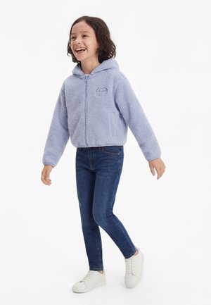 Lachend kind loopt, draagt een lichtpaarse fleece hoodie, donkerblauwe jeans en witte sneakers tegen een witte achtergrond.