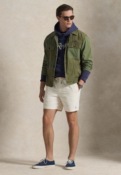 Μπουφάν ελιάς με μπροστινές τσέπες, μπλε-ναυτικό φούτερ με κουκούλα, λευκά σορτς με κορδόνια και μπλε αθλητικά παπούτσια. Αξεσουάρ περιλαμβάνουν γυαλιά ηλίου και ρολόι.