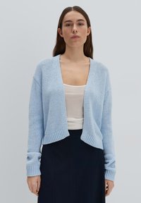 Cardigan tricoté bleu clair avec une finition légèrement texturée, devant ouvert et manches longues, associé à un haut blanc et une jupe foncée.