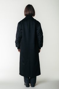 Casaco longo preto com gola, combinando tecido liso no corpo e material acolchoado nas mangas; design elegante e minimalista.