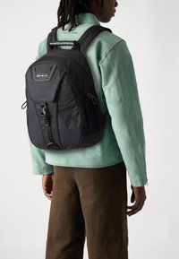 Mochila negra con una correa ajustable, bolsillos con cremallera y bolsillos laterales de malla. Tejido texturizado y diseño elegante. Llevada sobre una chaqueta verde clara.