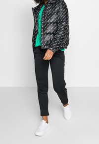 Chaqueta acolchada negra con acabado brillante y logo repetido de "DKNY", combinada con un sudadera verde y pantalones joggers negros, complementada con zapatillas blancas.