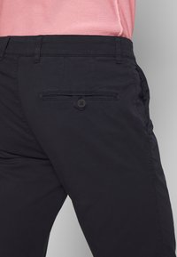 Ciemnogranatowe spodnie z tylną kieszenią na guzik, o płaskim kroju i gładkiej fakturze. Część różowej koszuli jest widoczna.