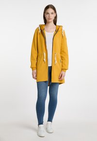 Giacca a vento gialla con zip frontale, dettagli con coulisse e due tasche laterali. Abbinata a jeans blu e sneakers bianche.