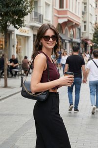 Jonge vrouw met zonnebril en een bordeauxrode mouwloze top draagt een zwarte handtas en houdt een koffiebeker vast op een drukke stadsstraat.