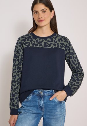 Kvinde iført en navy sweatshirt med grøn og sort leopardprint på ærmer og bryst, kombineret med blå denimjeans, stående med hånden i lommen.