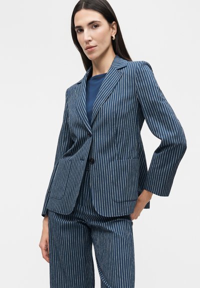 Γυναίκα που φοράει μπλε σκούρο σακάκι με ρίγες και ταιριαστό παντελόνι, με μπλε σκούρο τοπ από μέσα και χρυσούς κρίκους στα αυτιά.