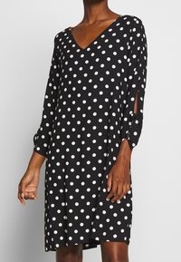 Vestido negro con lunares blancos, escote en V, mangas largas con aberturas, a la altura de la rodilla, tela suave con un diseño fluidos.