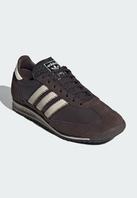 Crne i sive Adidas tenisice s gornjim dijelom od antilopa i kože, tri svijetlosive pruge sa strane i crne vezice na bijeloj pozadini.