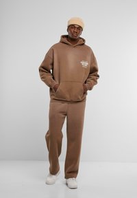 Sudadera marrón y pantalones a juego hechos de material suave, con un bolsillo frontal; presenta un logo en el pecho y un gorro crema.